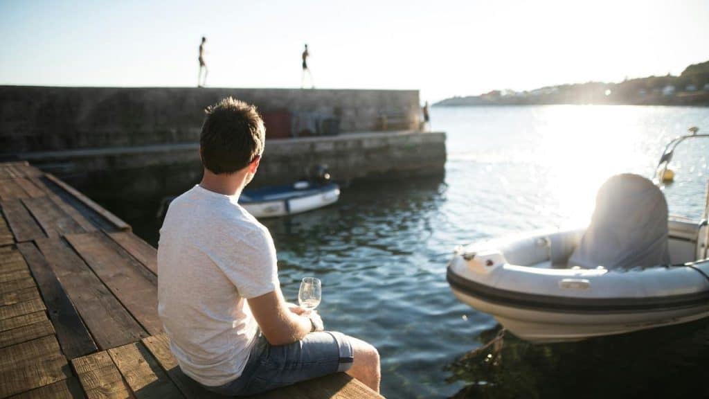 A man sitting on a dock holding a wine glass and looking at the water.