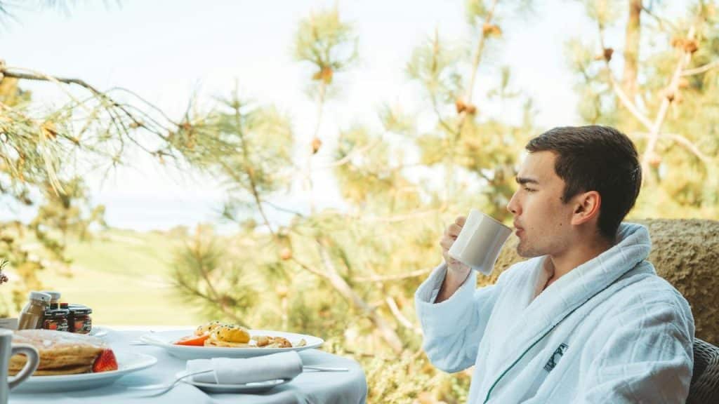 A man in a white robe drinking from a mug at an outdoor breakfast table.