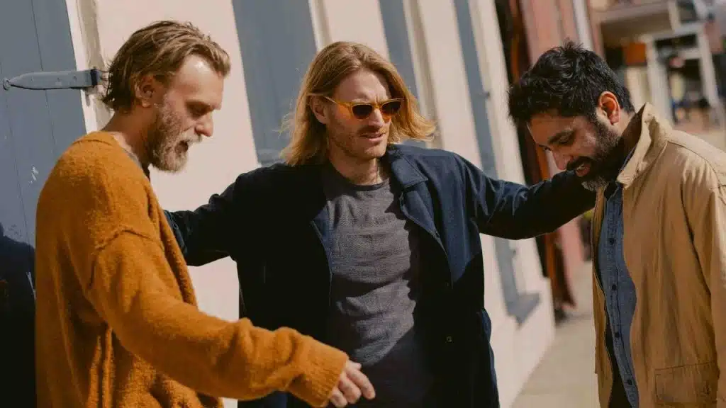 A man with sunglasses has his arms around two friends while walking outside.
