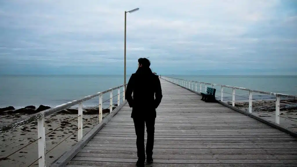 A man in a dark coat walking alone on a pier over the ocean.