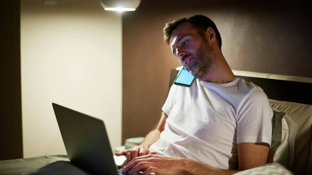 A man lies in bed using a laptop while holding a phone to his ear.