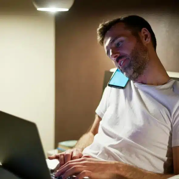 A man lies in bed using a laptop while holding a phone to his ear.