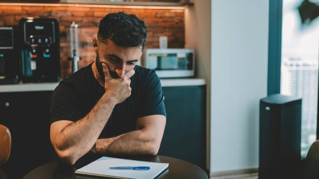A man sits at a table with his hand over his mouth, looking down at a notebook.