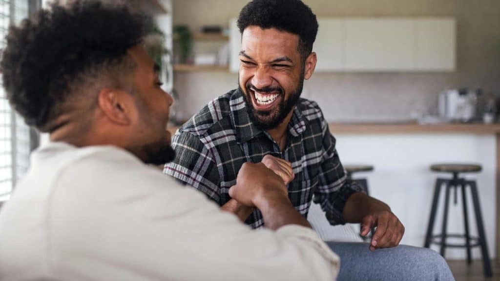 A pair of men laughing and shaking hands in a kitchen.
