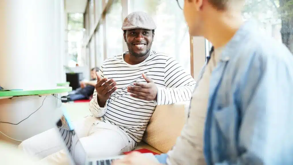 A man in a striped shirt and cap smiling while talking to another person indoors.