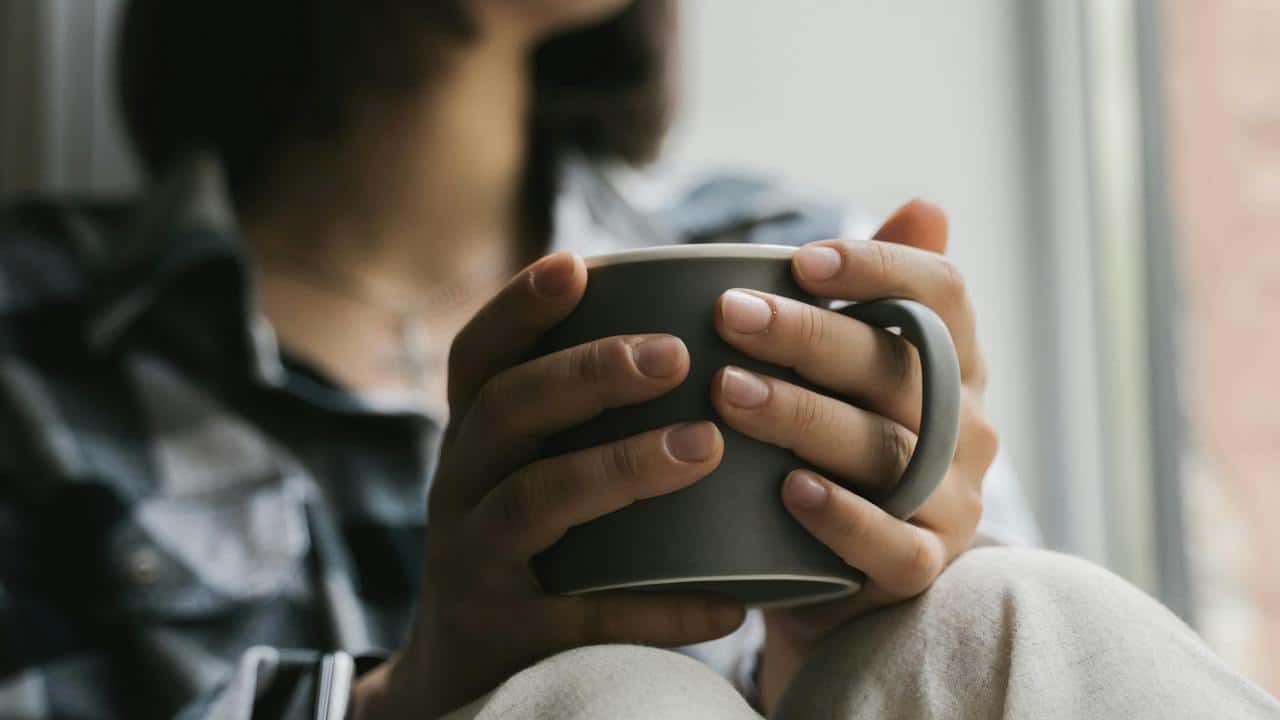 A person holds a mug with both hands while sitting by a window.