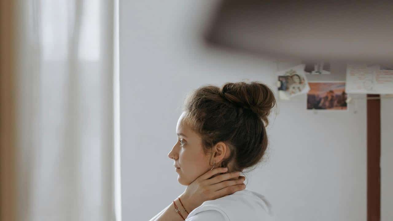 A woman sits indoors with her hand on her neck, looking pensive.