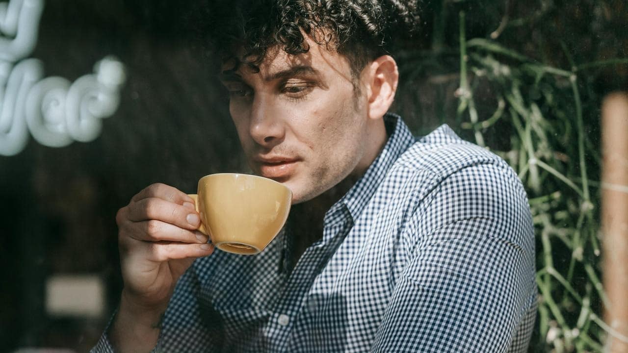A man in a checkered shirt sits by a window holding a yellow cup.