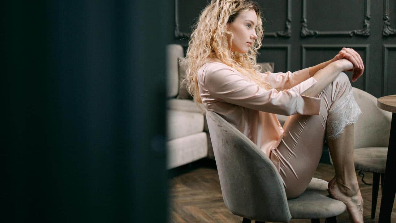 A woman in loungewear sits curled up on a chair, looking thoughtful.