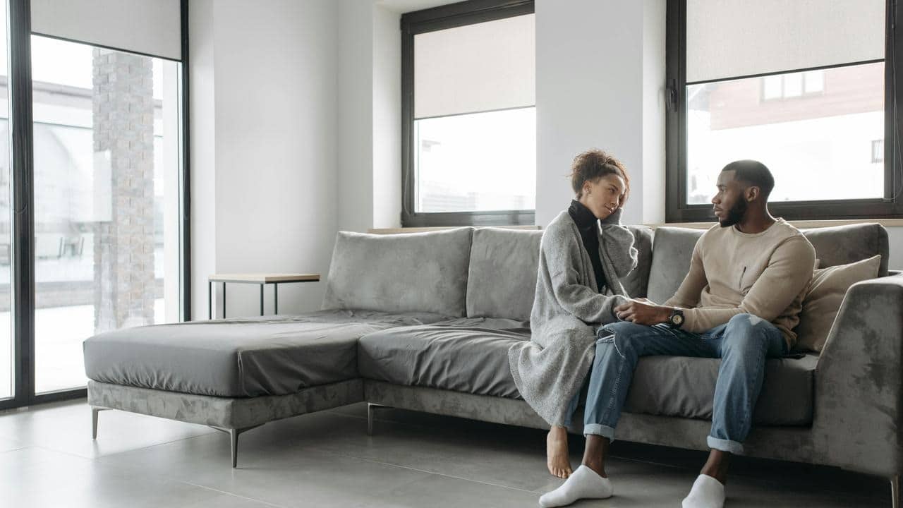A couple sits on a couch holding hands and talking.
