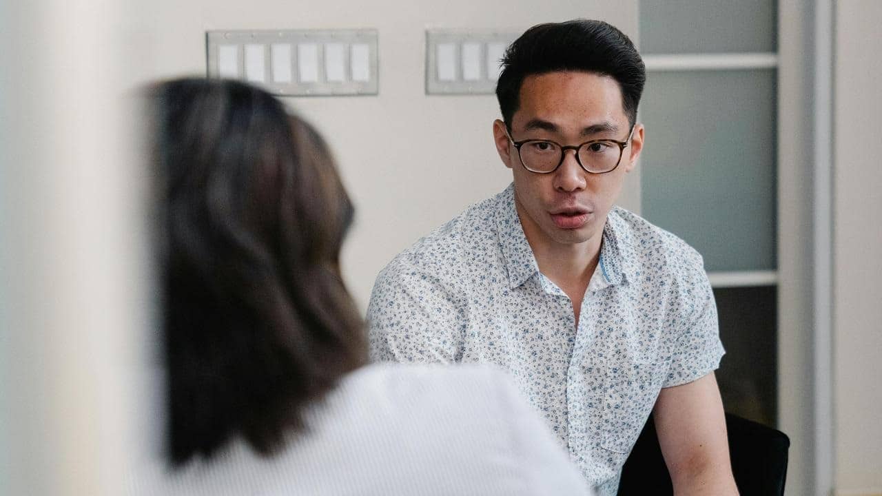 A man wearing glasses talks to someone in a room.