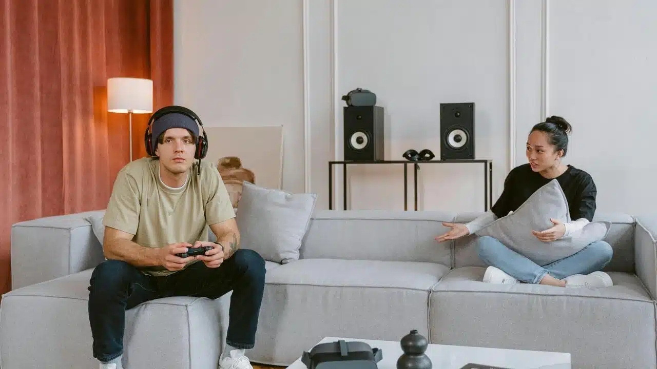 A man gaming while a woman speaks to him on the couch.