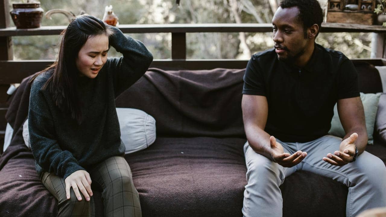 A couple sitting outside on a couch, with the woman upset and the man talking to her.