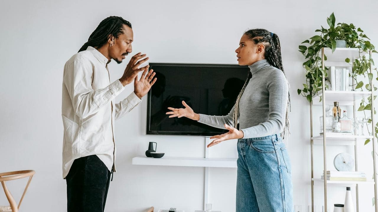 A couple standing indoors, facing each other and arguing with expressive gestures.
