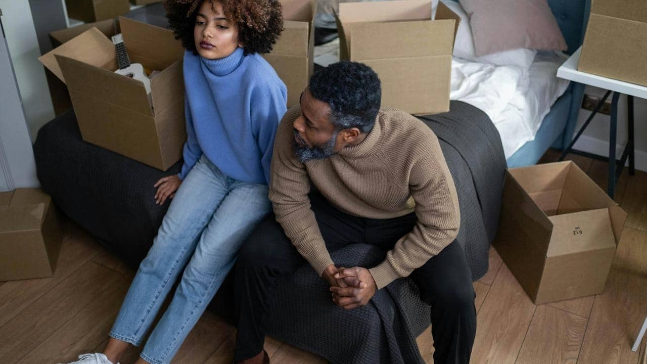 A couple sitting on a bed surrounded by moving boxes, both looking upset and distant.
