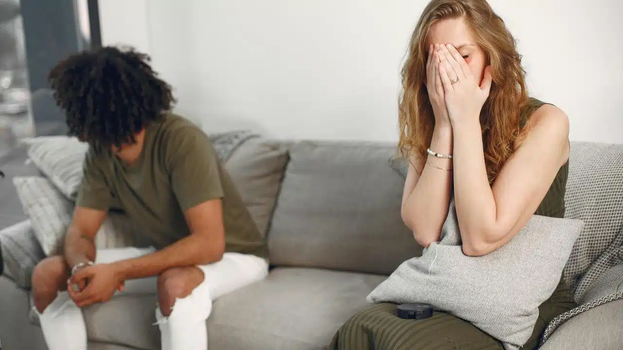 A woman covering her face on a couch while a man sits beside her facing away.