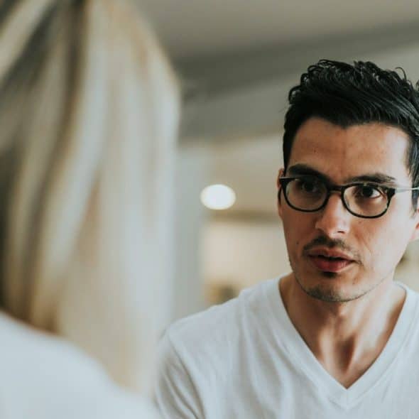 A man wearing glasses looks intently at a woman during a serious conversation in a bright indoor setting.