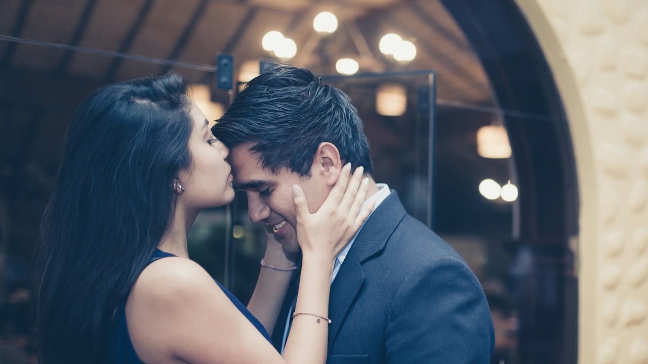 A woman in a blue dress gently kisses a man in a suit on the forehead.