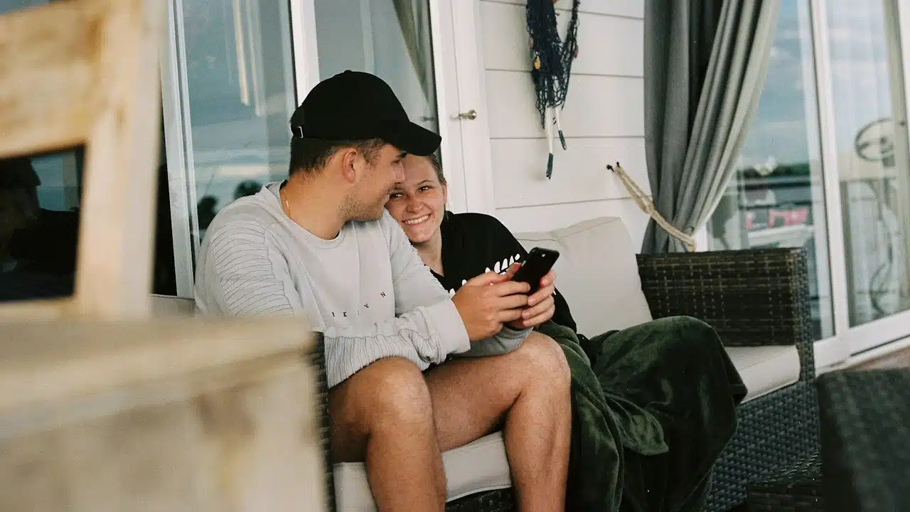 A smiling woman leaning against a man who is using his phone while they sit on a couch outdoors in front of a house.