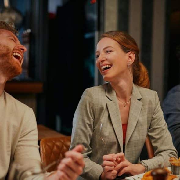 A joyful couple at dinner, with a red-haired man laughing heartily and a smiling woman in a blazer looking at him.