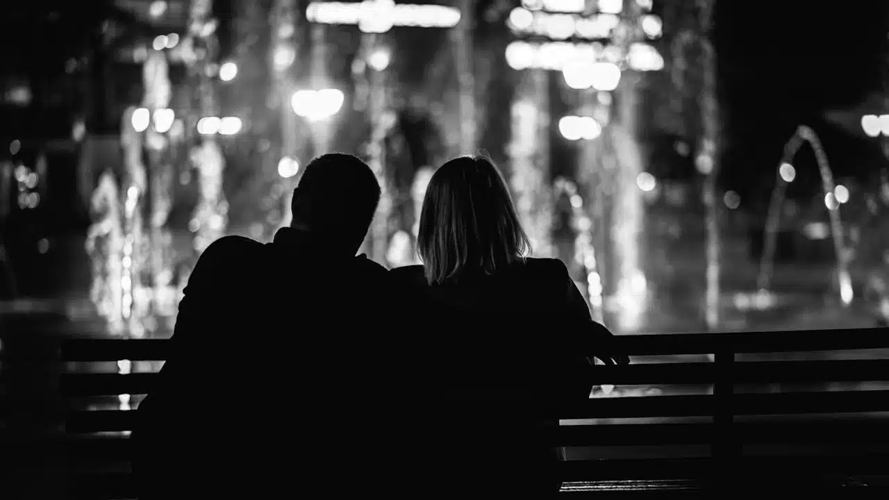 A black and white image of two people, viewed from the back, sitting on a bench and looking at illuminated fountains at night.