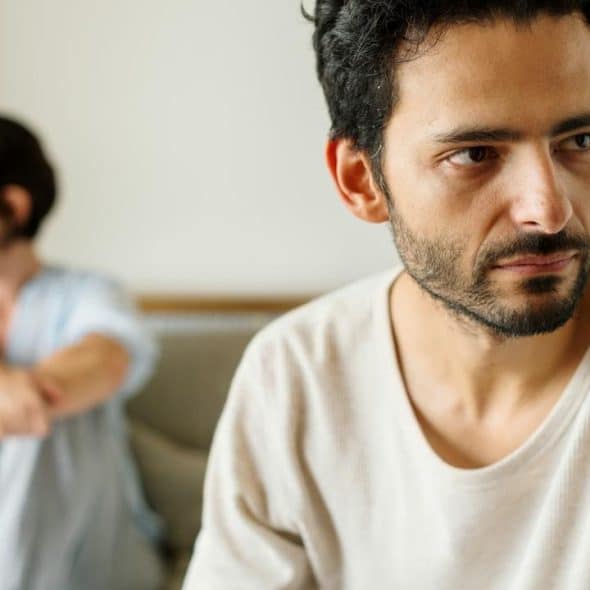 A man looks away with a tense expression while a woman sits blurred in the background.