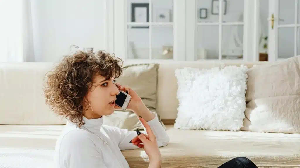 A woman sitting on the floor talking on her smartphone in a bright living room.