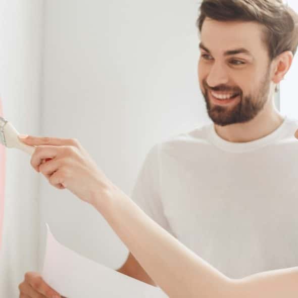 A smiling couple painting a wall together, with one person holding a paintbrush and the other holding a sheet of paper, as they work on a light-colored wall with pink paint.
