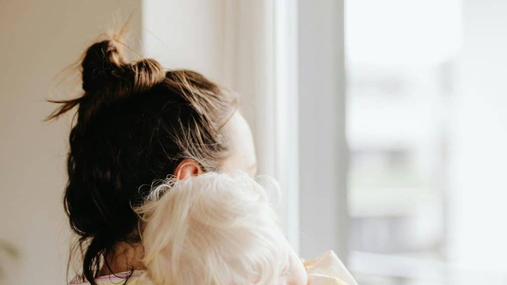 A woman holds a baby while looking out a bright window.