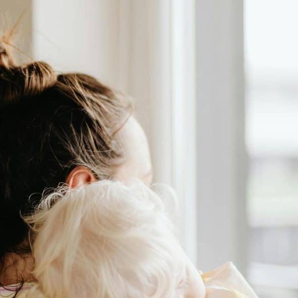 A woman holds a baby while looking out a bright window.