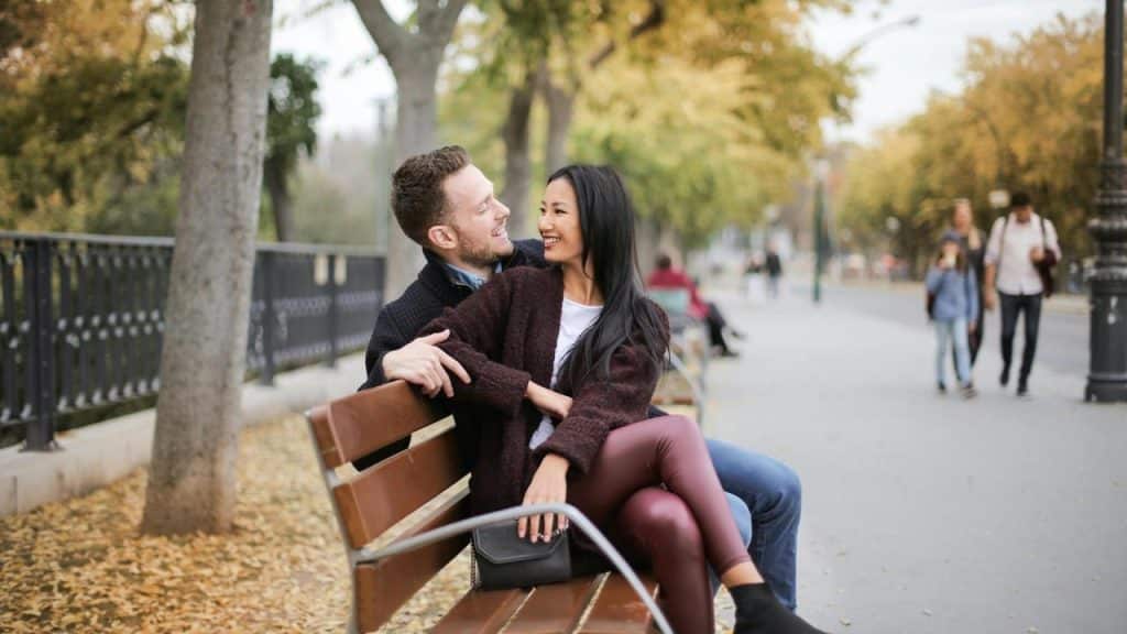 A couple sitting on a park bench smiling at each other.