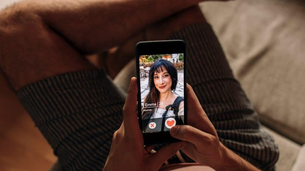 A person holding a smartphone showing a dating app profile of a woman, with options to like or reject the profile visible on the screen.