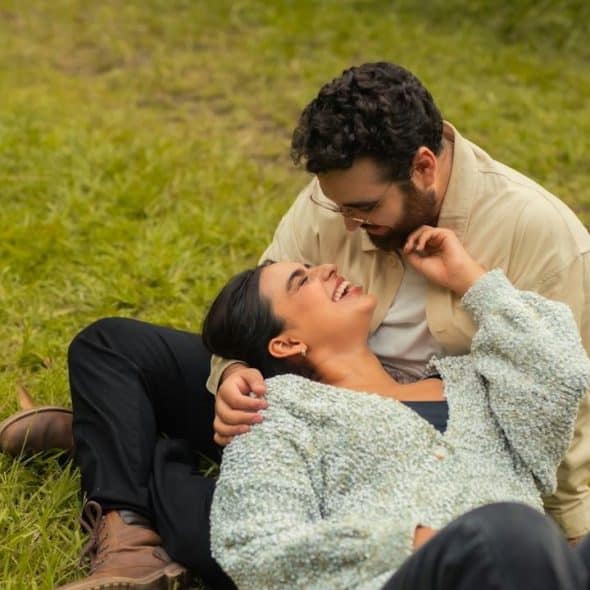 A happy couple outdoors, with the woman lying back on the grass and laughing, and the man leaning over her, smiling while touching her face.