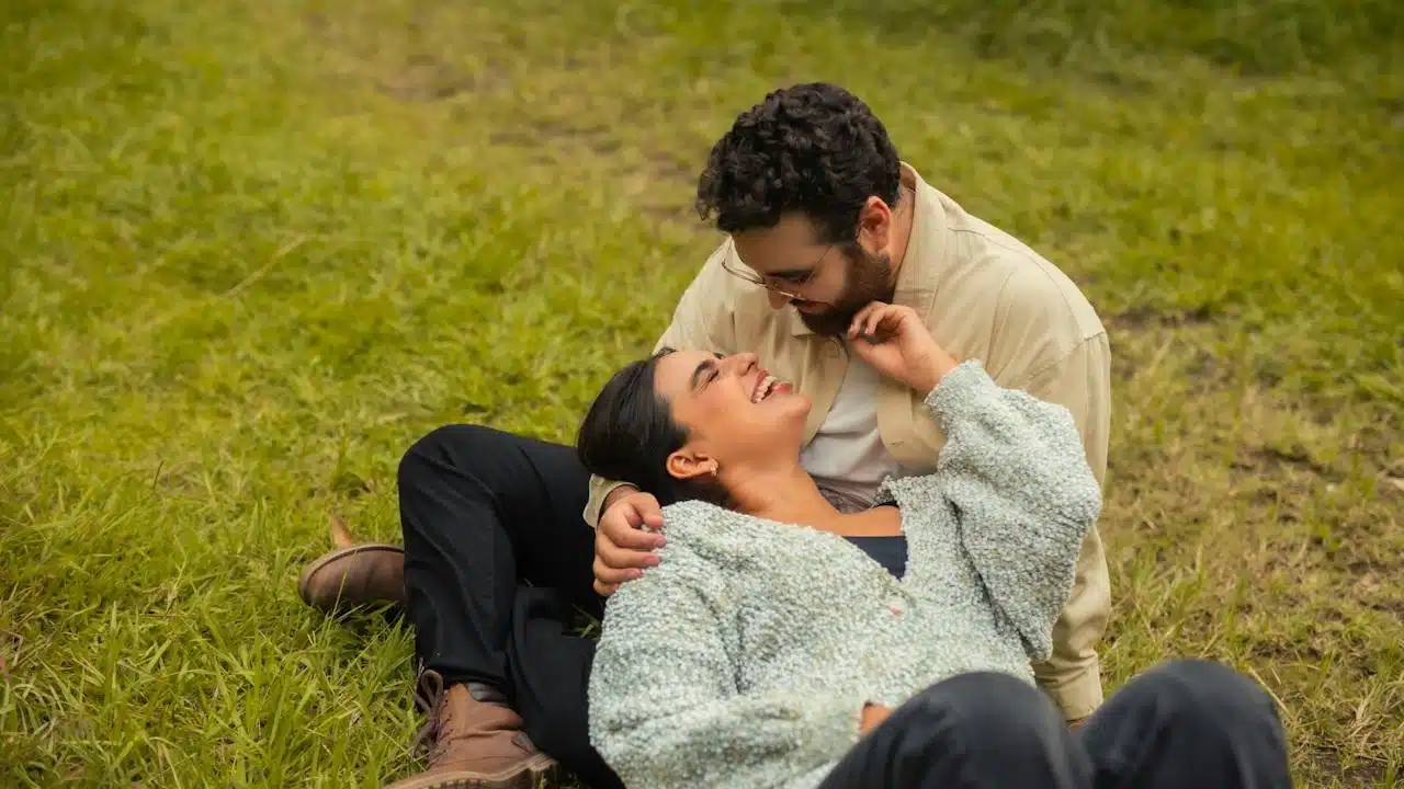 A happy couple outdoors, with the woman lying back on the grass and laughing, and the man leaning over her, smiling while touching her face.