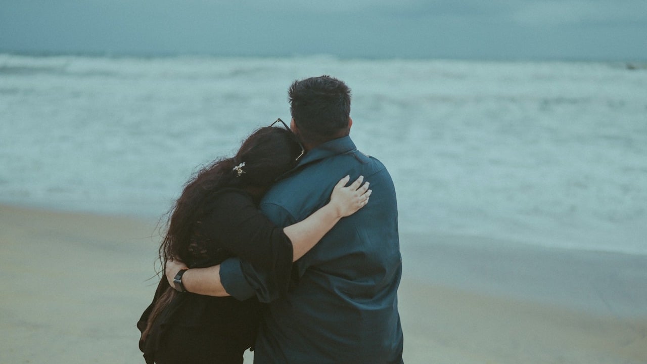 A rearview shot of a couple standing on a sandy beach, looking out at the ocean waves under an overcast sky.