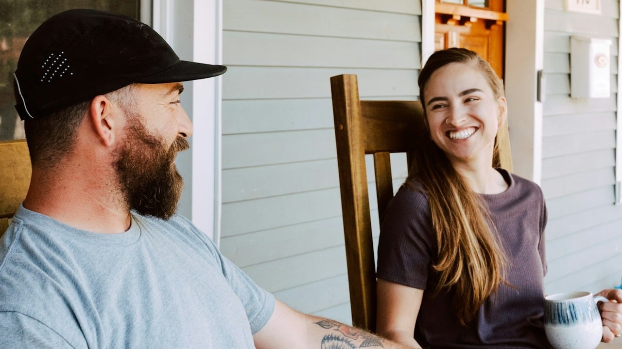 A bearded man wearing a cap and a smiling woman sit on a porch, facing each other and chatting.