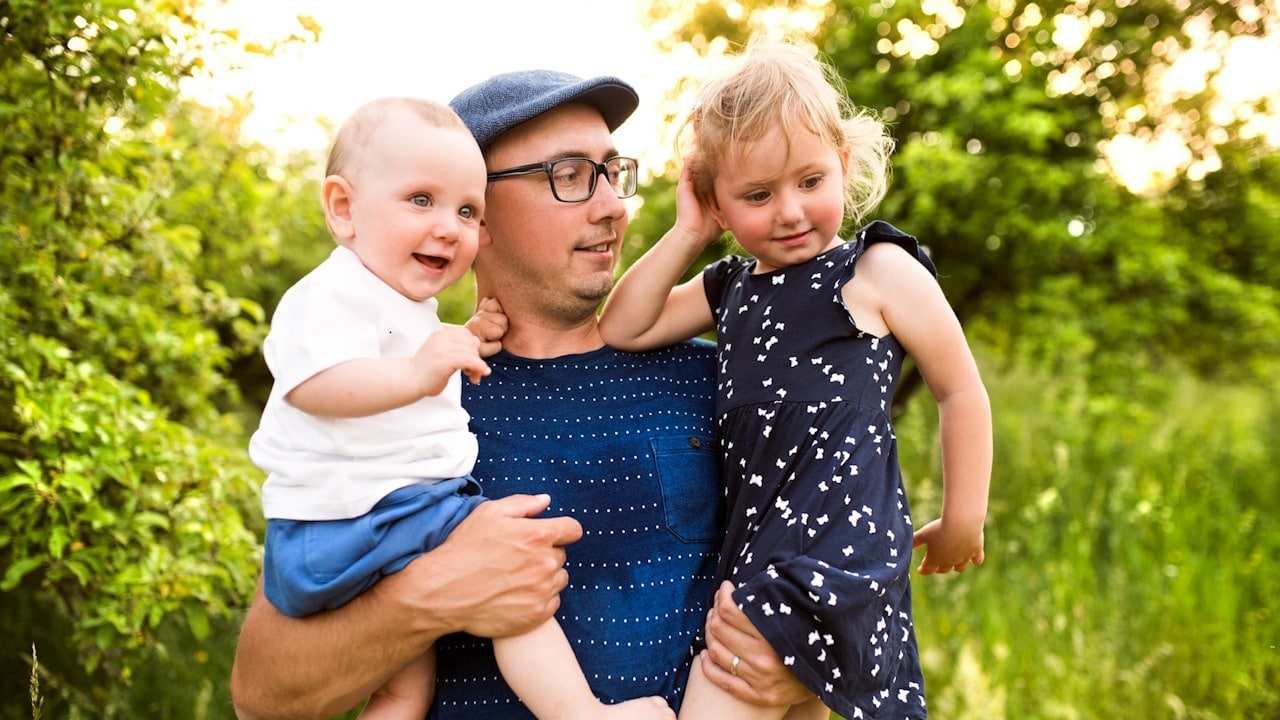 A man wearing a cap and wearing glasses holding his kids while standing outdoors.