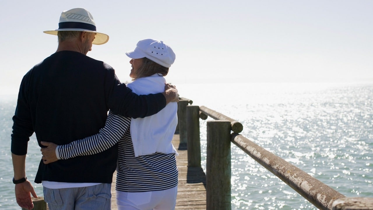 A couple walking down a pier with their arms around each other’s backs and smiling at each other.