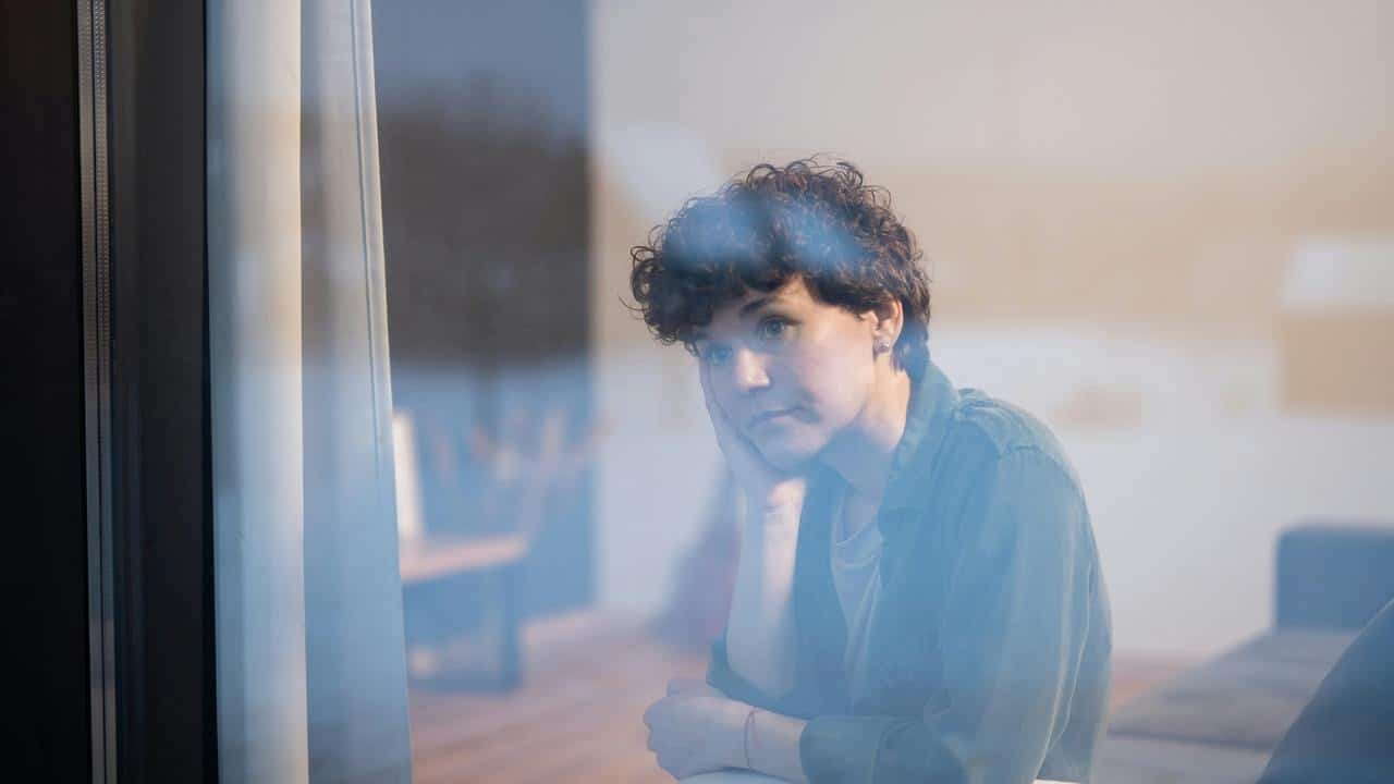 A person sitting indoors and gazing out a window with a thoughtful expression.