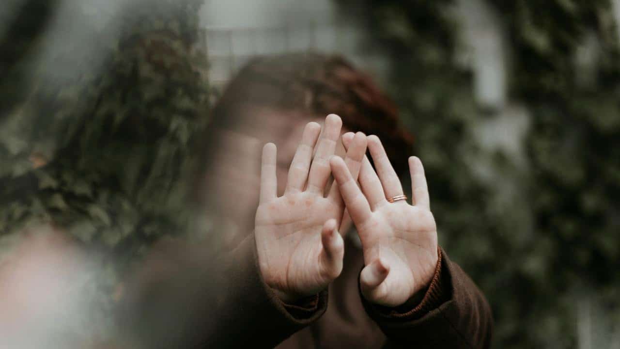 A person holding up their hands to cover their face behind glass.