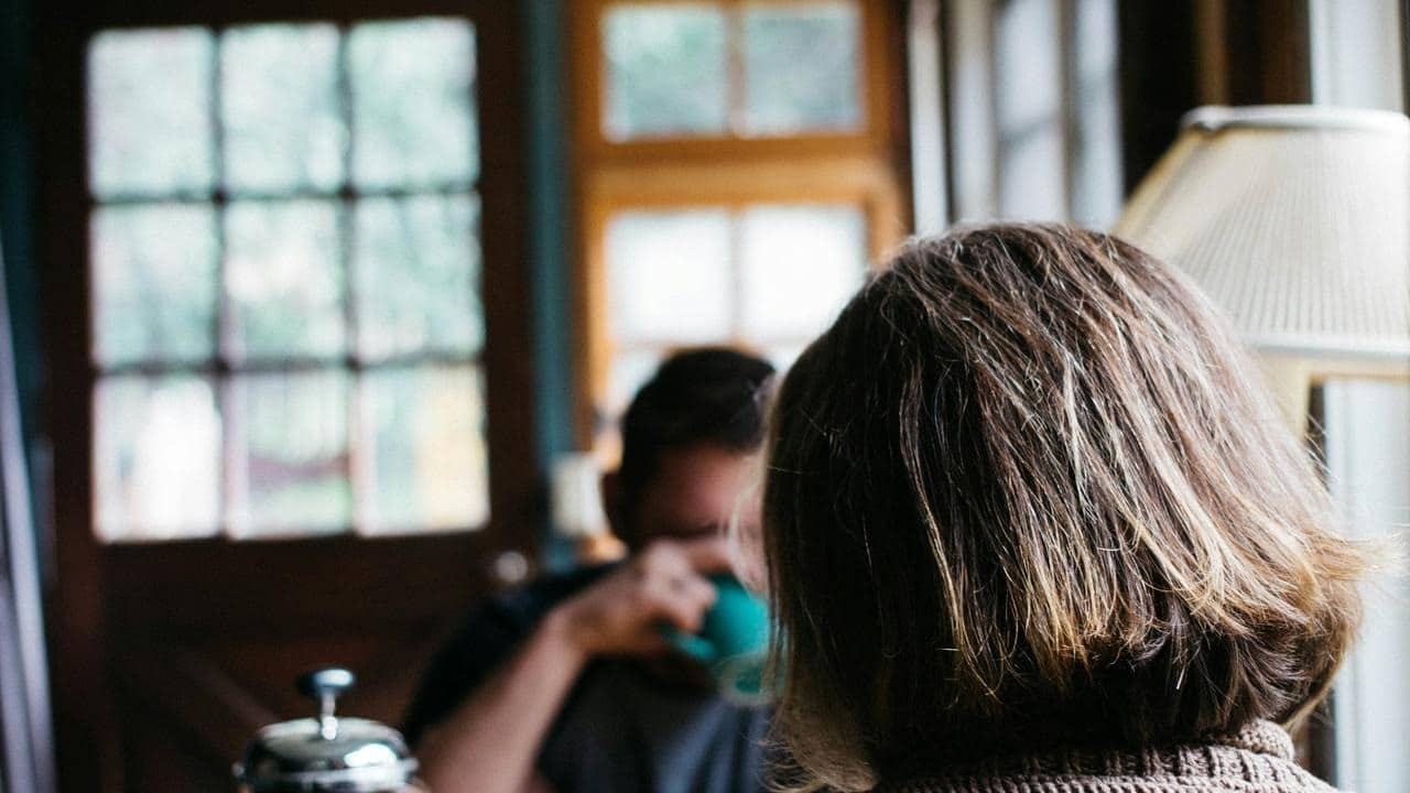 A person sitting indoors with another person blurred in the background.