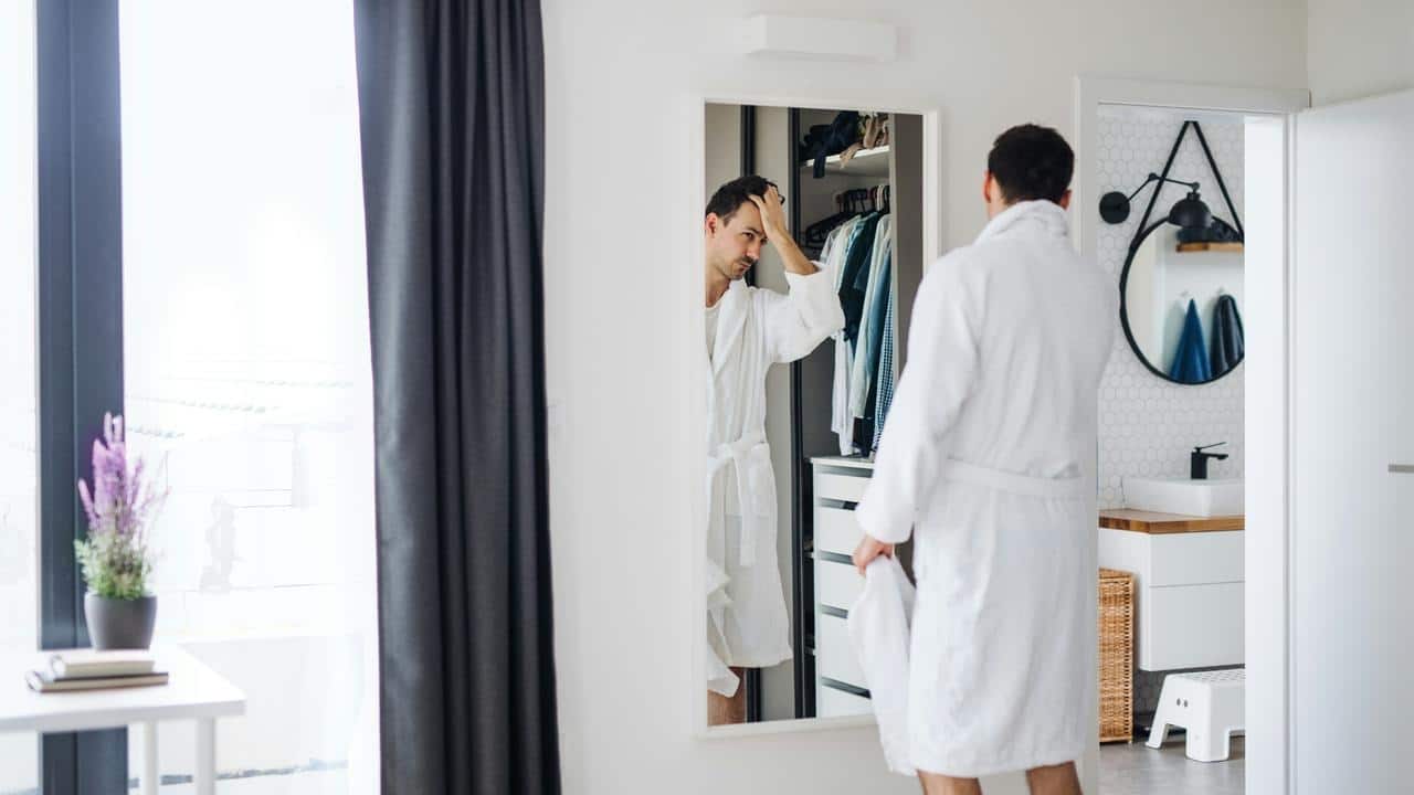 A man in a bathrobe looking at himself in a mirror in a bedroom.