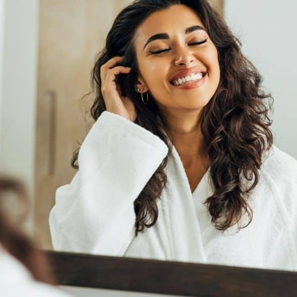 A smiling woman in a white robe looking at herself in a mirror.