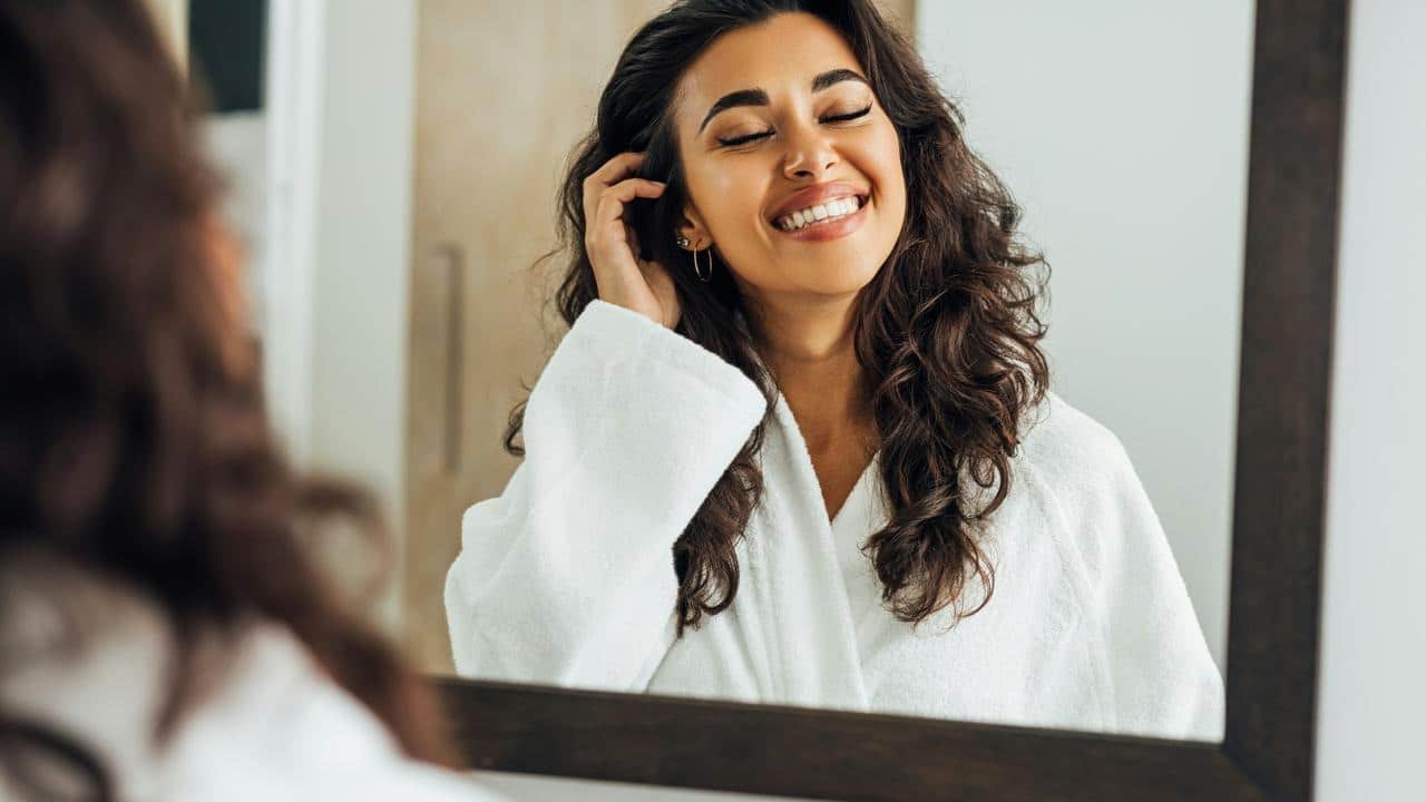 A smiling woman in a white robe looking at herself in a mirror.