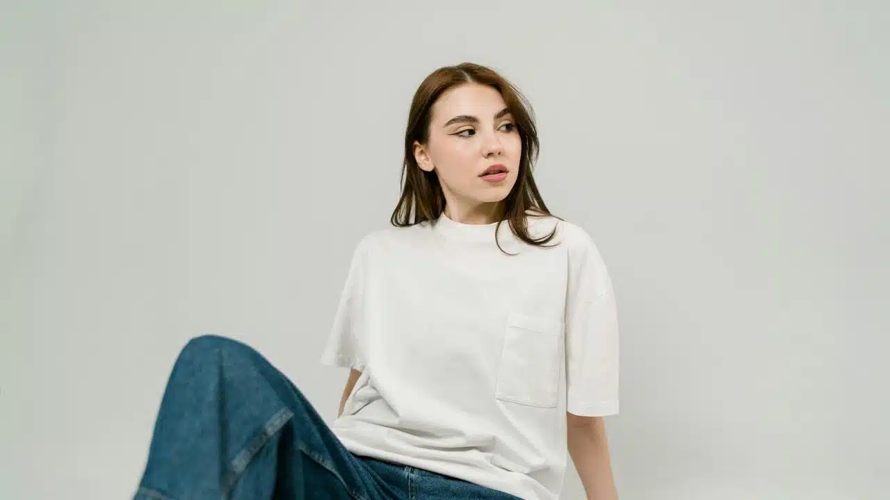 A woman in a white shirt sits and looks to the side against a plain background.