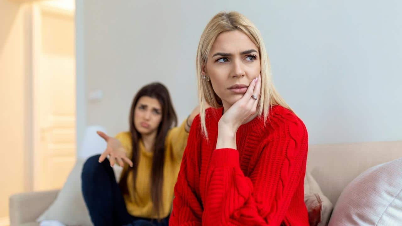 A woman in a red sweater looks upset while another woman gestures in the background.
