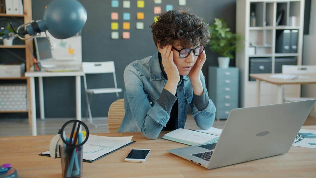 A stressed woman sits at a desk holding her head while looking at a laptop.
