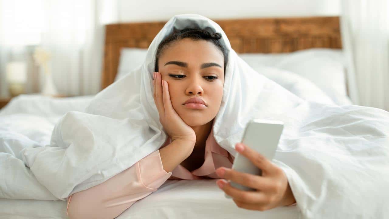 A woman lies in bed under a blanket while looking at her phone.