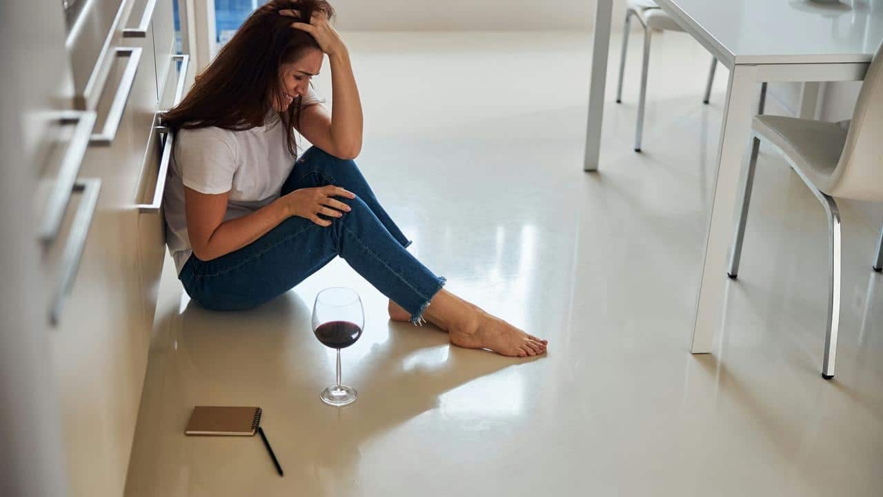 A distressed woman sits on the floor with a glass of wine nearby.