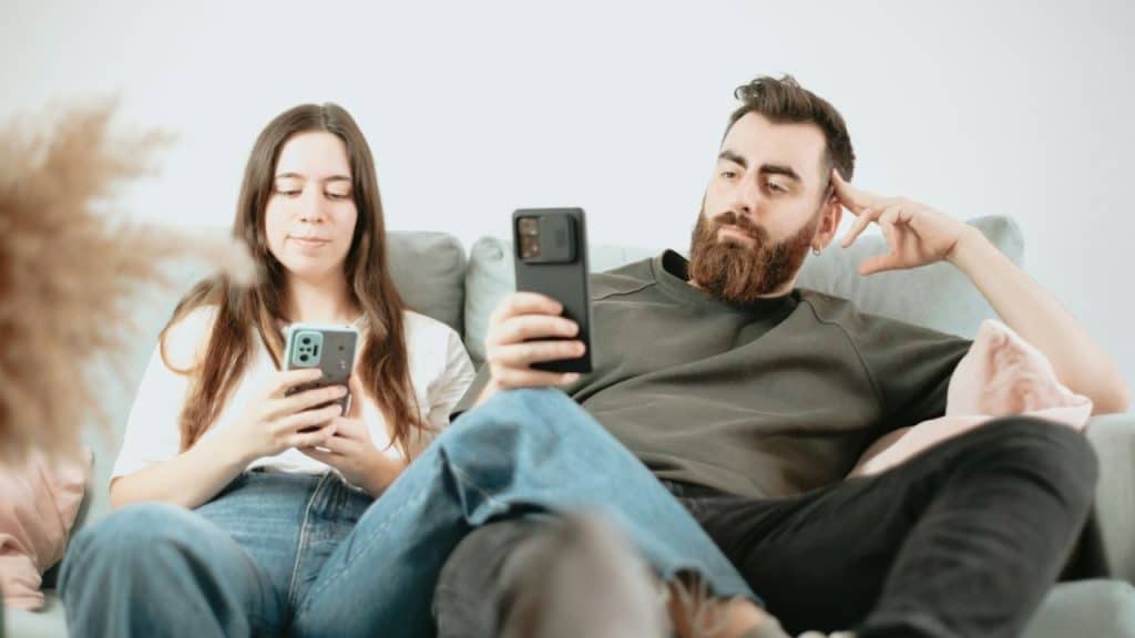A man and a woman sit together on a couch while both look at their smartphones.